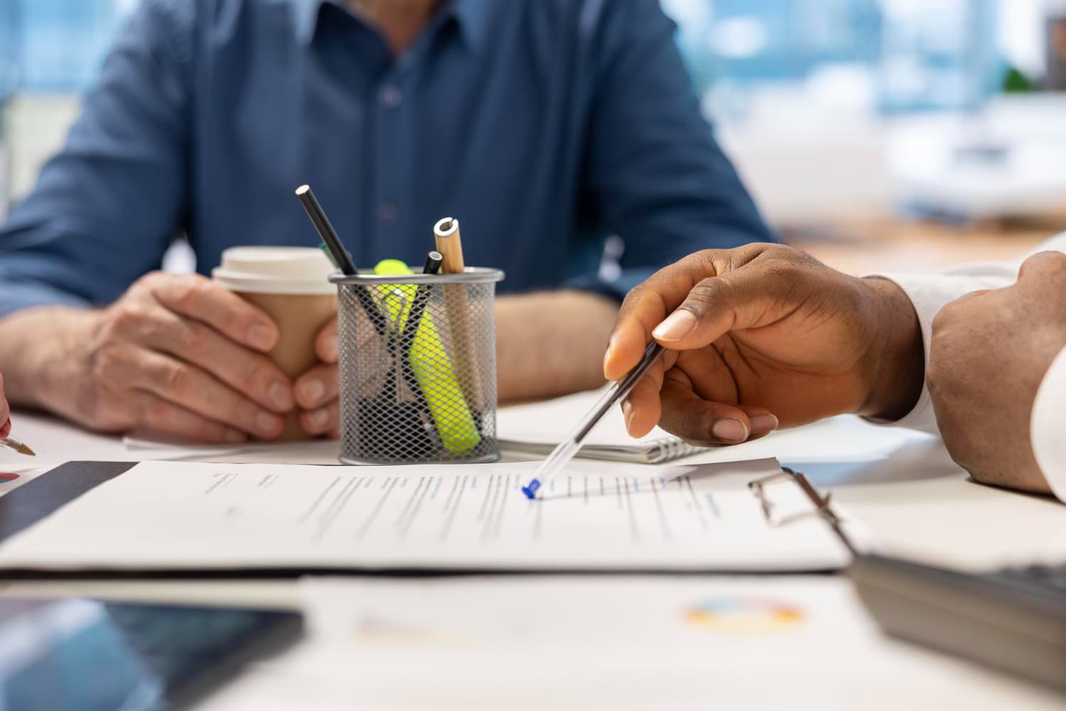 Two colleagues at a desk going over a printed form with a pen, beside a coffee cup and a pot of pens